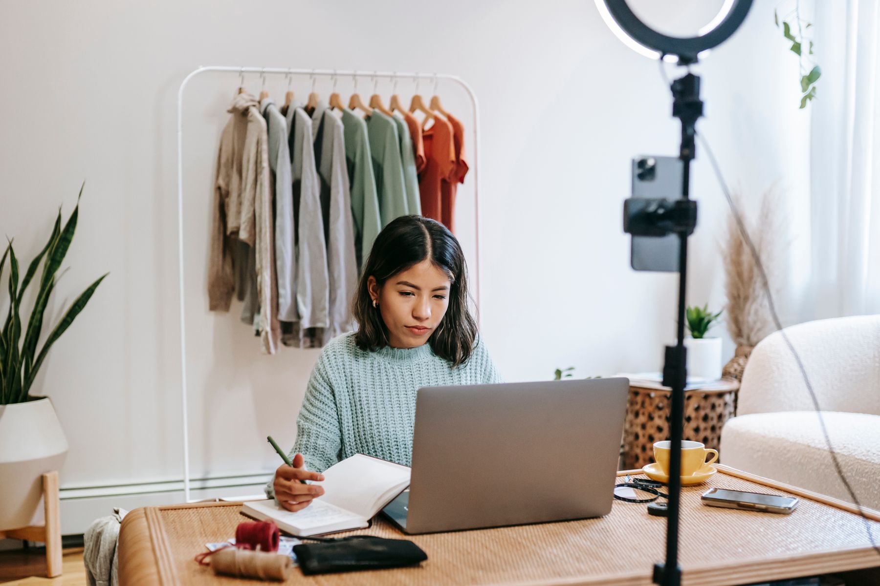 A creator works on her laptop and notebook beside a recording setup in a bright home studio.