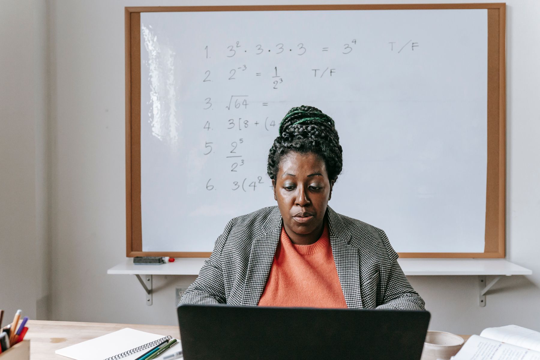 An educator teaches online from a laptop with a mathematics whiteboard behind her.